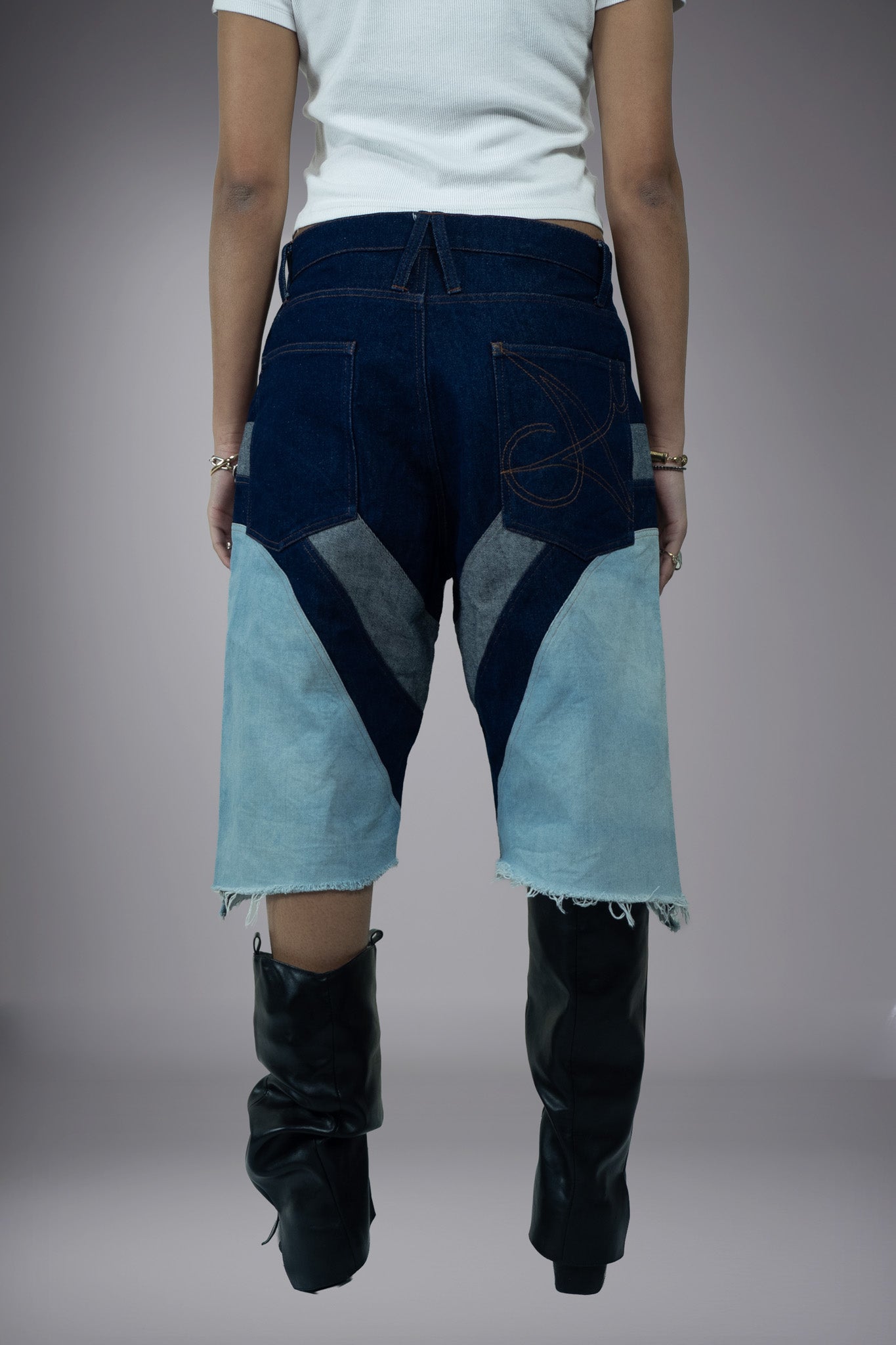 ack view of two-tone denim long shorts with dark raw denim pockets, light wash leg panels, graphic pocket stitching and raw frayed hems.