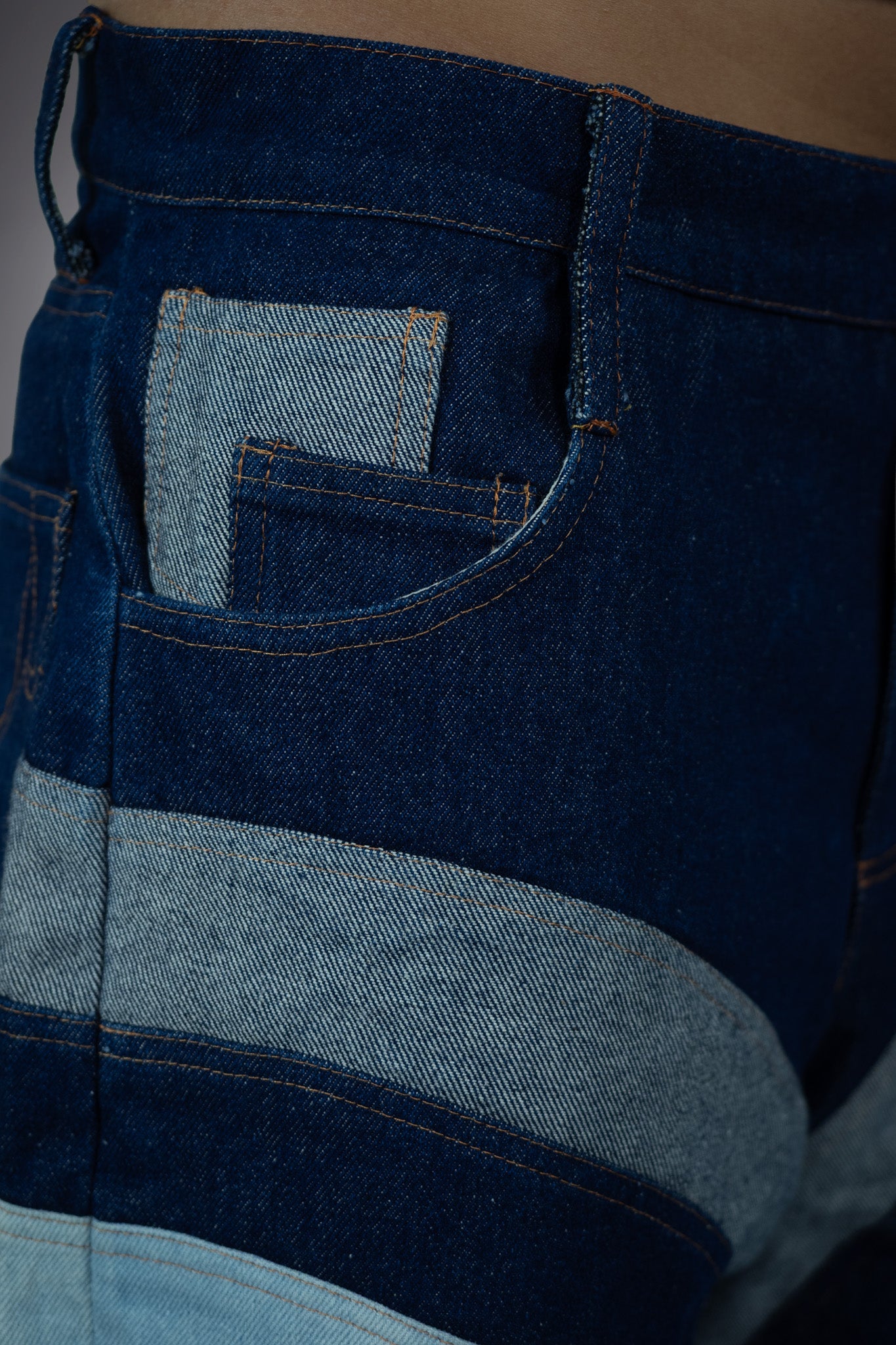 Close-up side pocket detail of two-tone denim long shorts with dark raw denim, light wash pocket insert and curved panel construction.