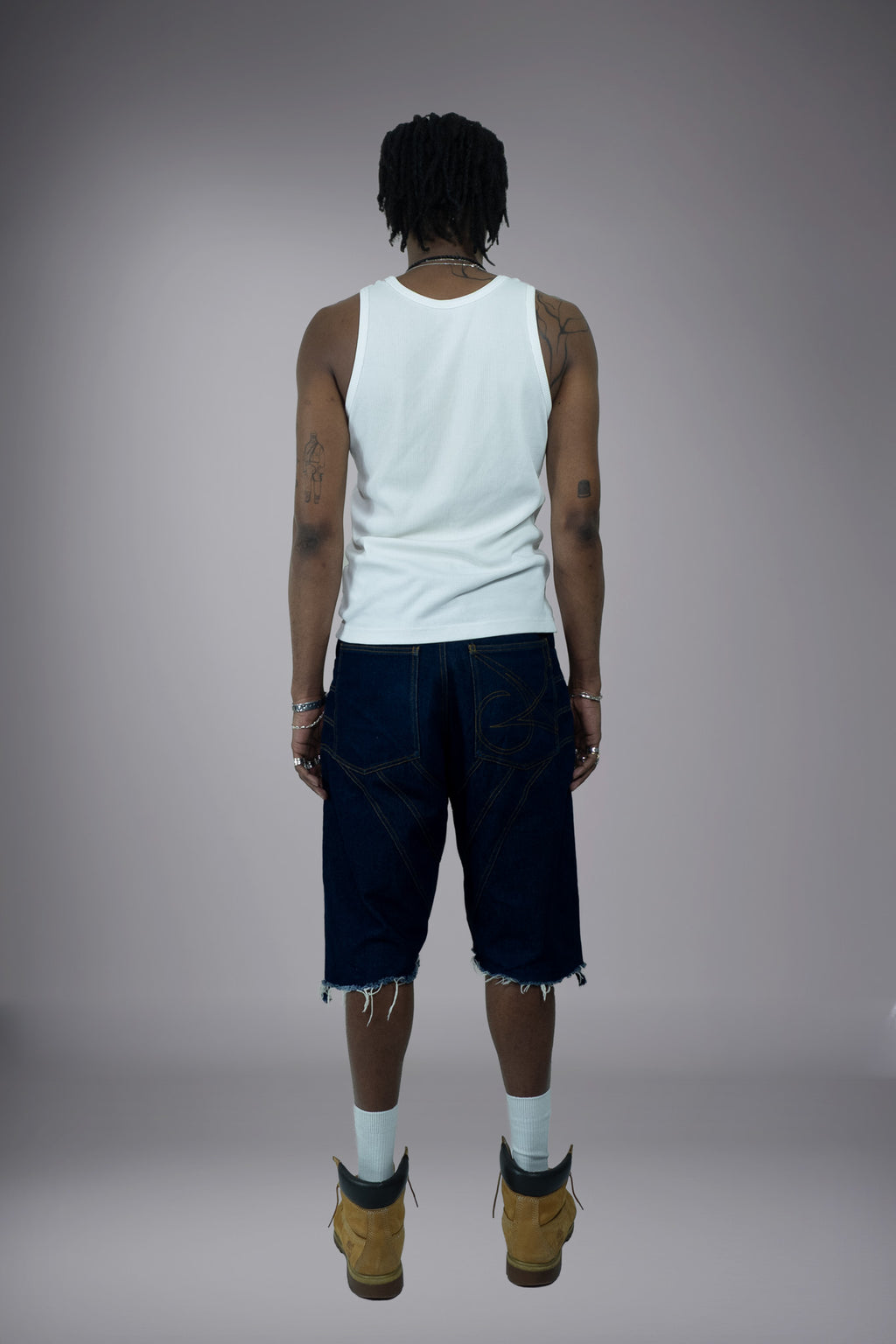 Back view of dark raw denim long shorts with graphic pocket stitching, wide-leg fit and raw frayed hem.