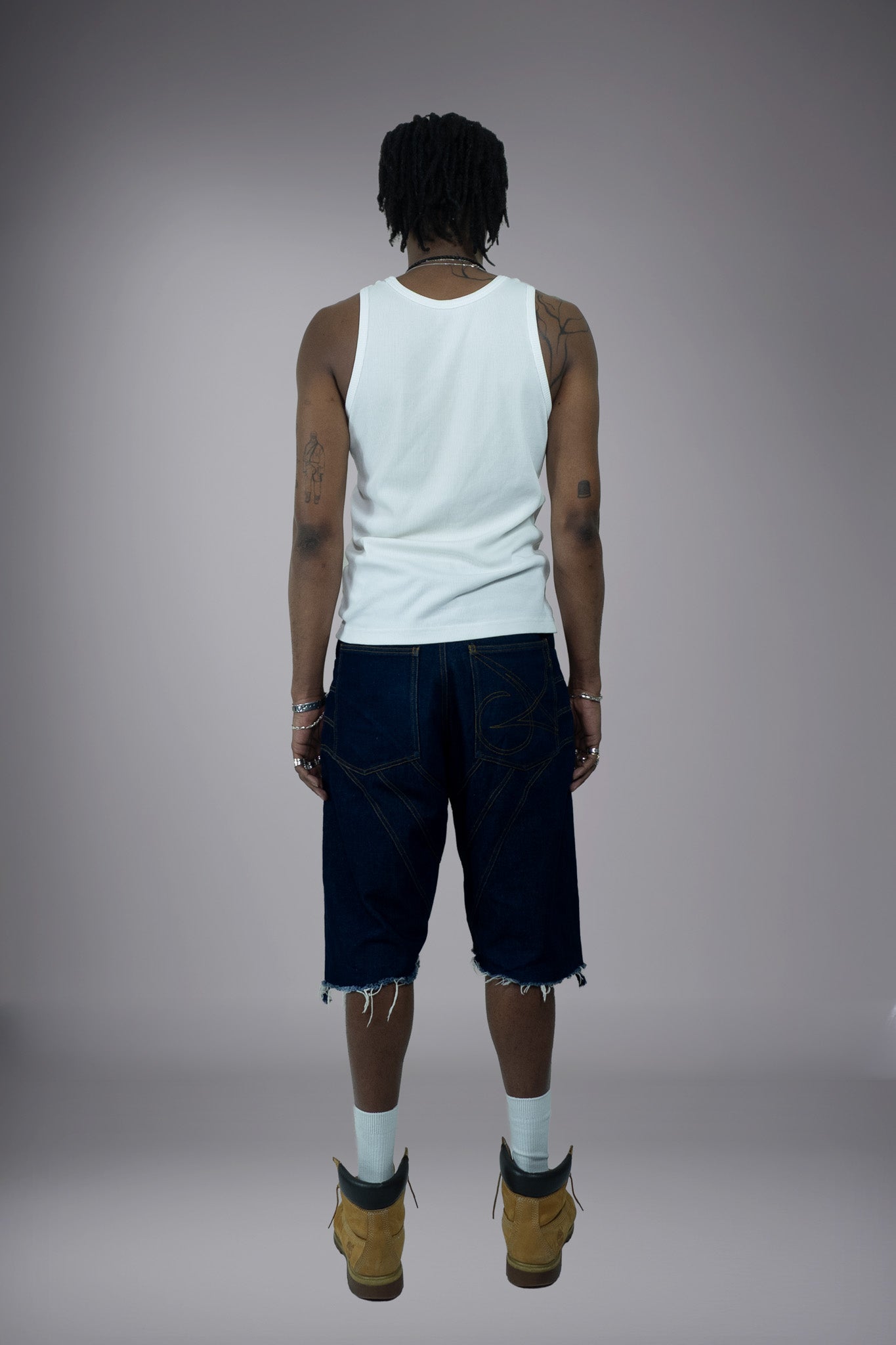 Back view of dark raw denim long shorts with graphic pocket stitching, wide-leg fit and raw frayed hem.
