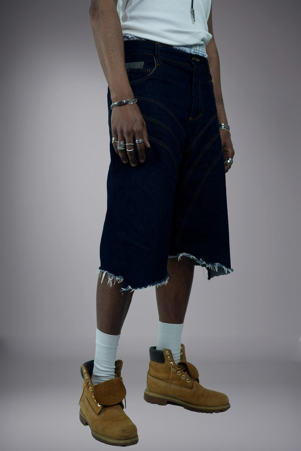 Close-up front view of dark raw denim long shorts with curved contrast stitching, wide-leg cut and raw frayed hem.