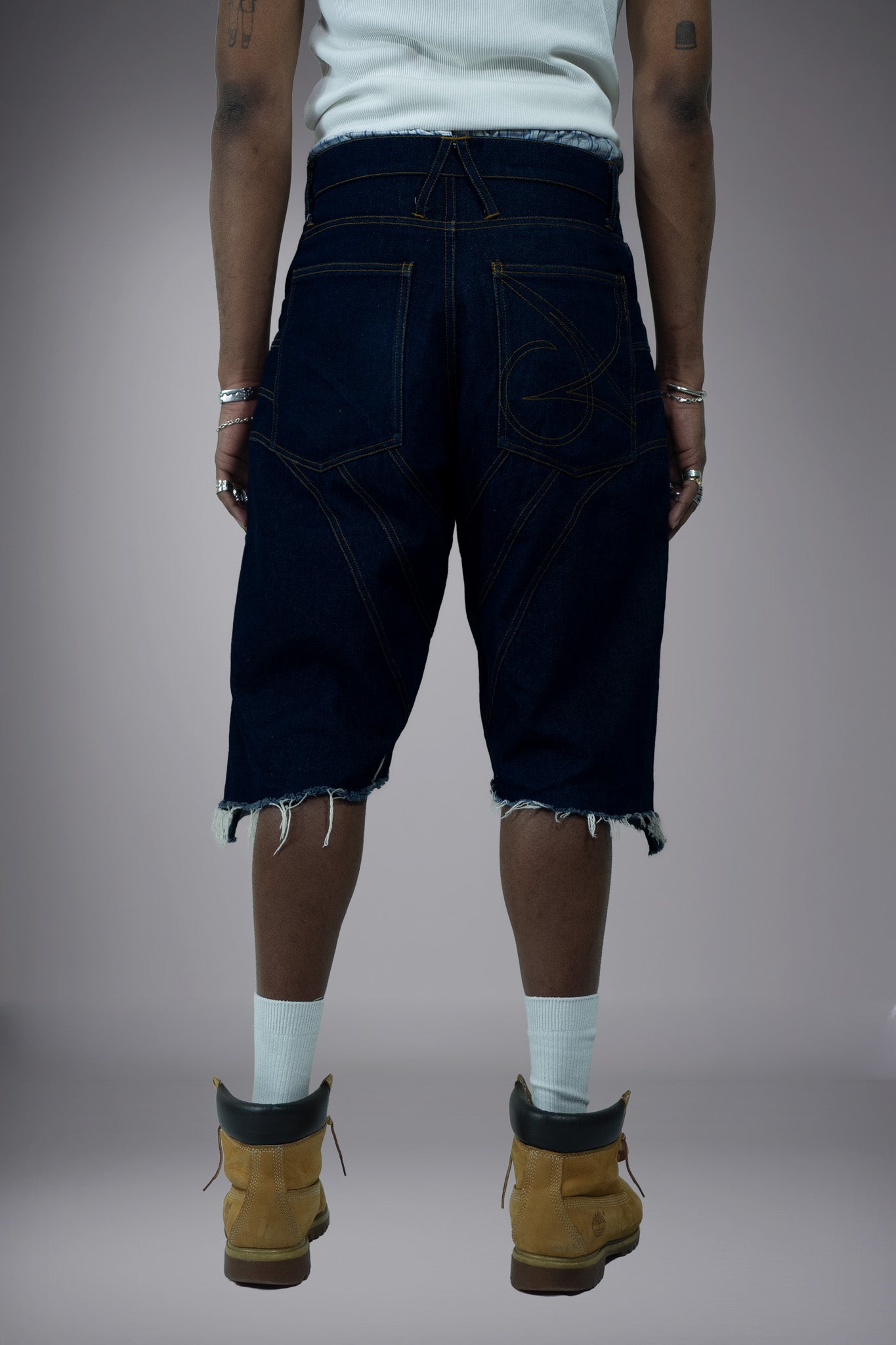 Back view of dark raw denim long shorts with graphic pocket stitching, curved contrast seams and raw frayed hems.