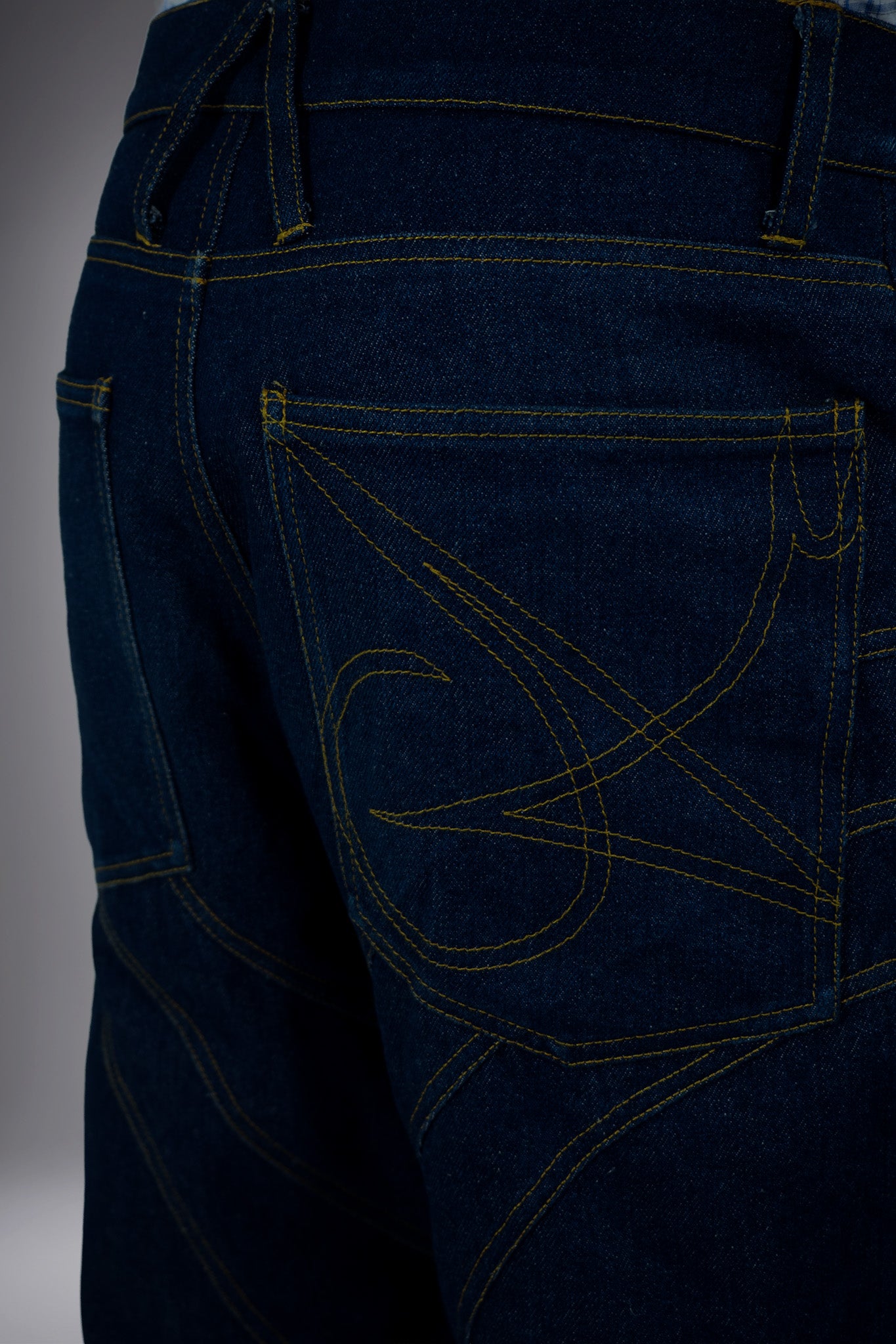 Close-up back detail of dark raw denim long shorts with graphic pocket stitching, contrast seams and structured waistband.