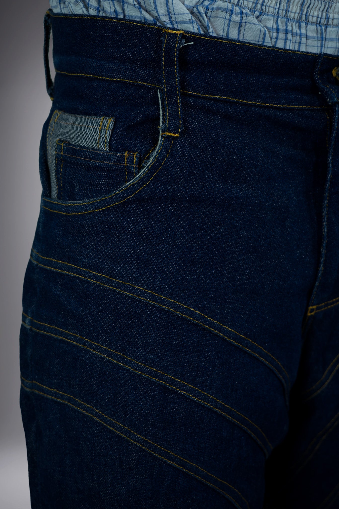 Close-up front detail of dark raw denim long shorts with curved contrast stitching, coin pocket and structured waistband.