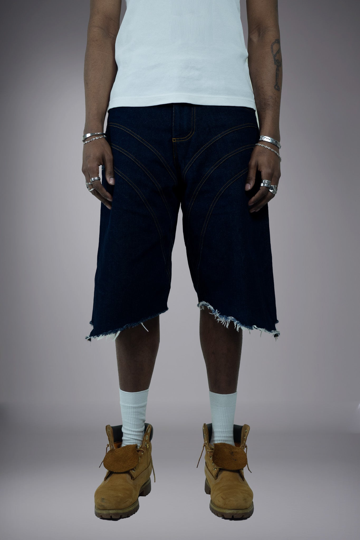 Front view of dark raw denim long shorts with curved contrast seam lines, wide-leg fit and raw frayed hem.